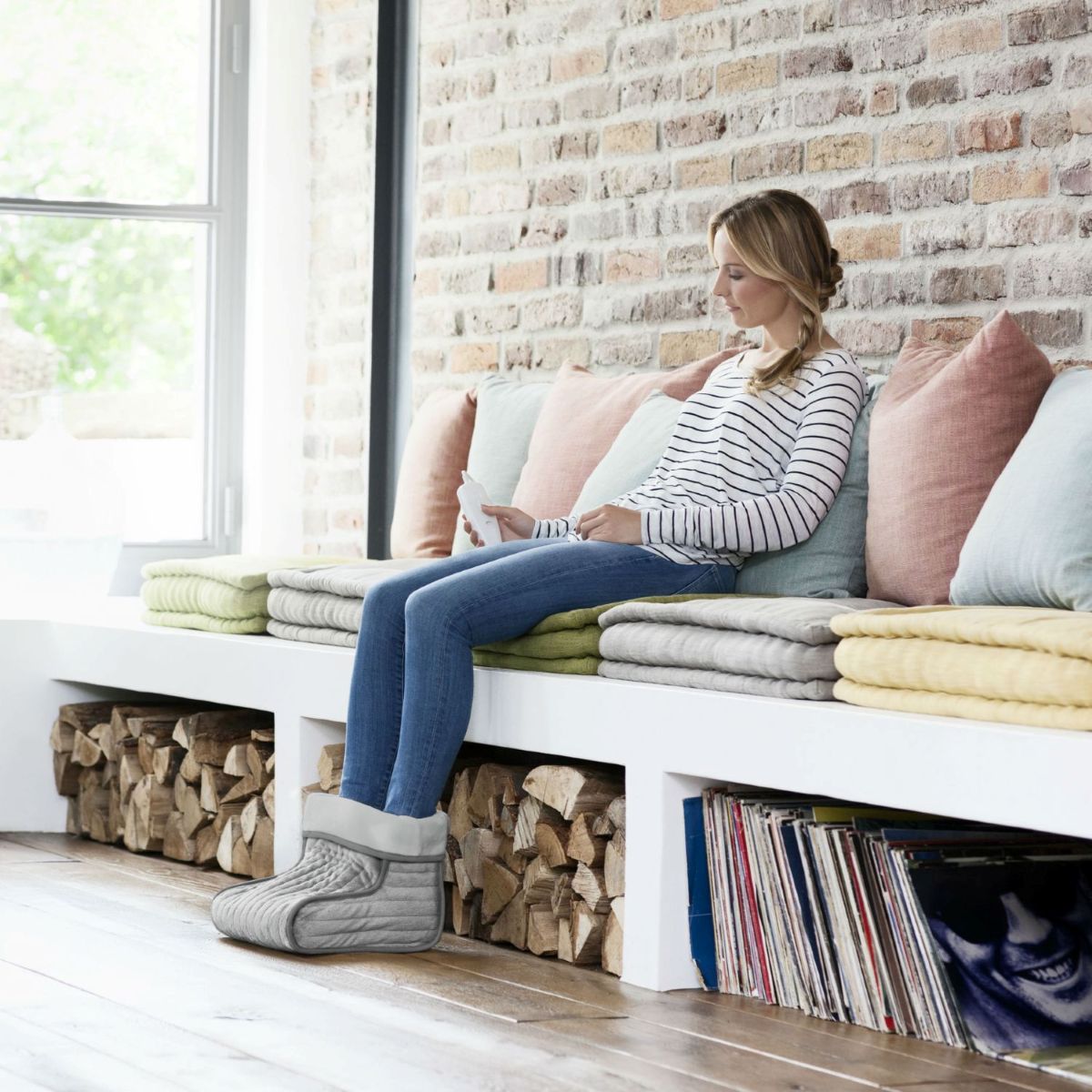 A woman relaxes on a cushioned bench wearing a grey electric foot warmer while holding a small device.